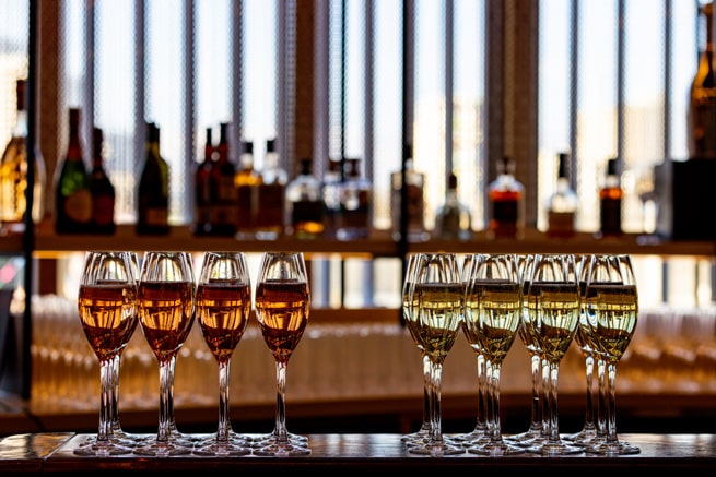 A group of flute glasses filled with sparkling wines. A bar shelf filled with various spirits is in the blurred background